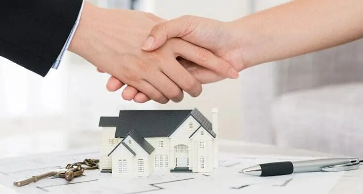 Two people shaking hands over a model house.